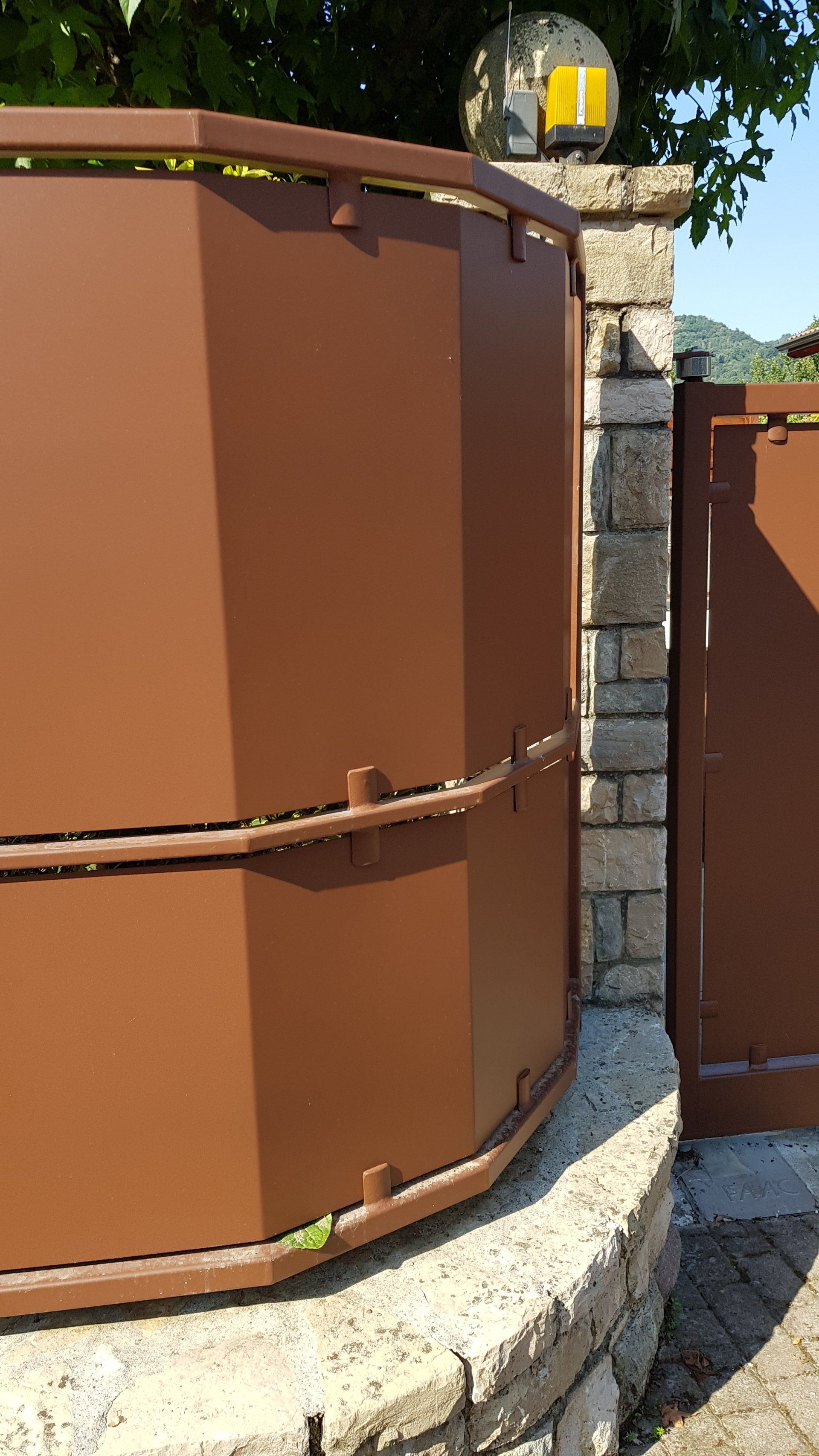 Cancello in metallo marrone accanto a un pilastro e a un muro in pietra, in un ambiente esterno soleggiato.