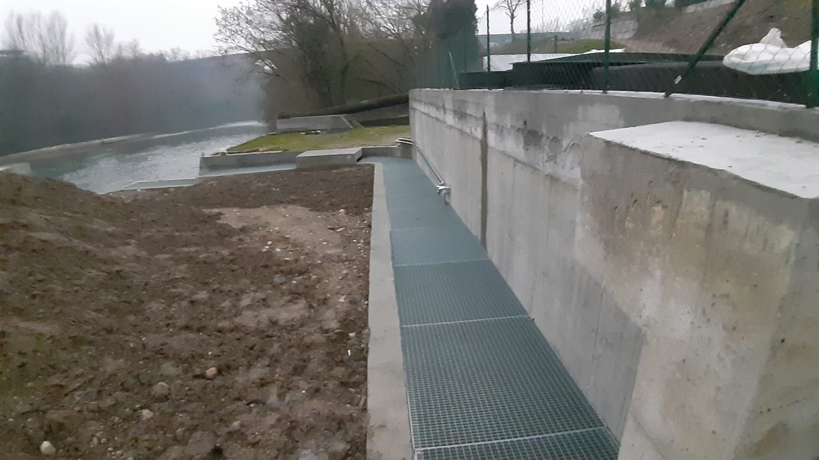 Passerella in cemento lungo il fiume, con sentiero in griglia metallica e muro di contenimento.
