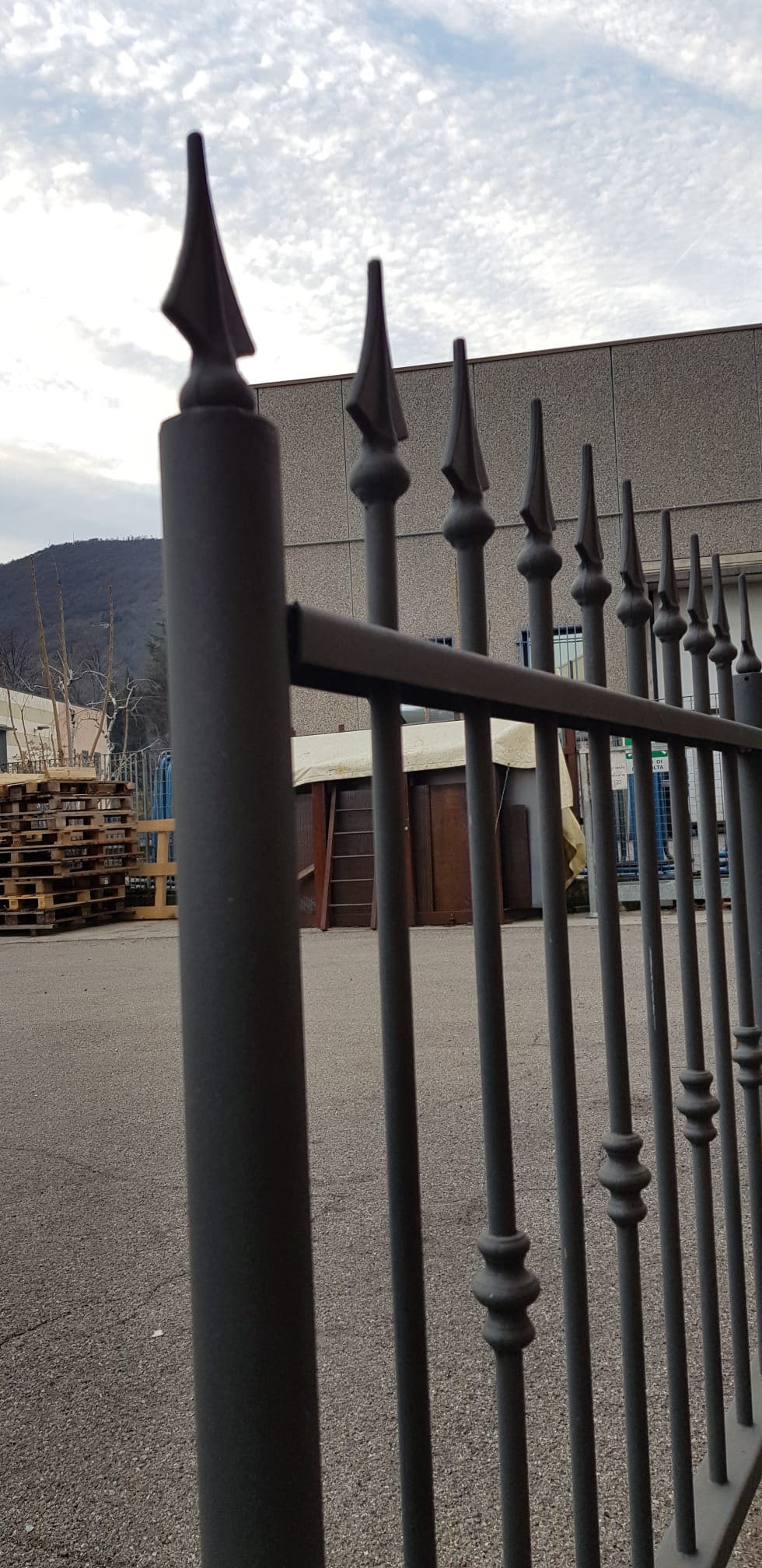 Recinzione in ferro nero con pali appuntiti a forma di lancia accanto a un edificio sotto un cielo nuvoloso.