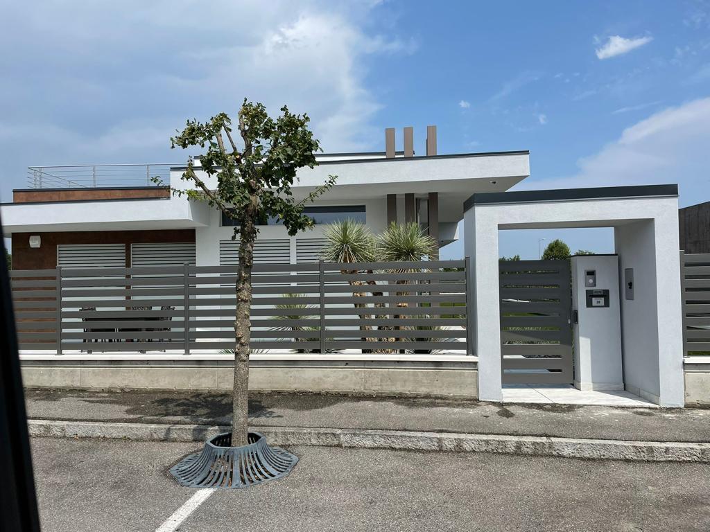 Casa moderna bianca con recinzione e cancello grigi e un giovane albero su una strada soleggiata