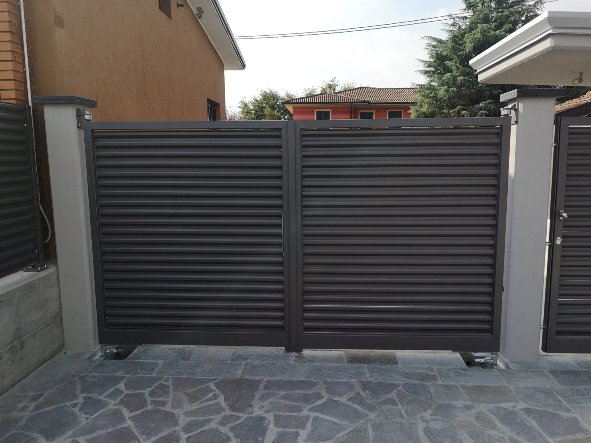 Cancello carrabile in metallo scuro con lamelle orizzontali tra muri in pietra e patio piastrellato