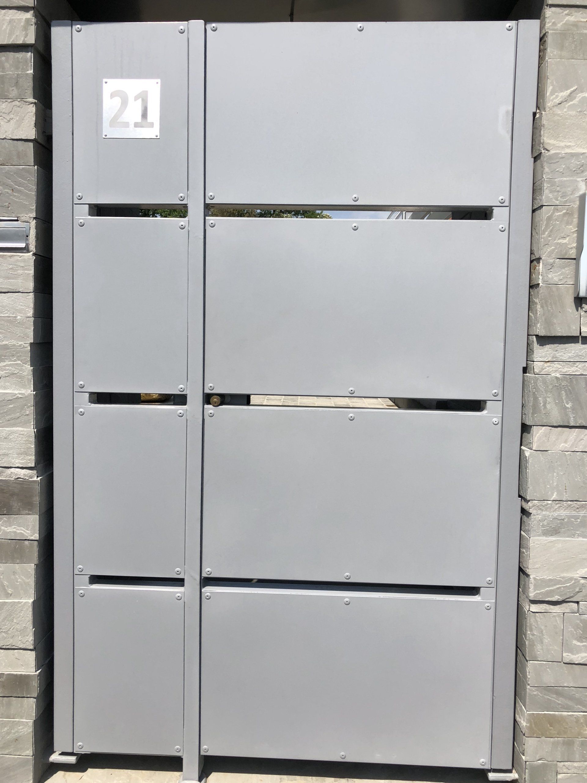 Cancello in metallo grigio con una piccola targa numerica e pannelli orizzontali su un muro di pietra