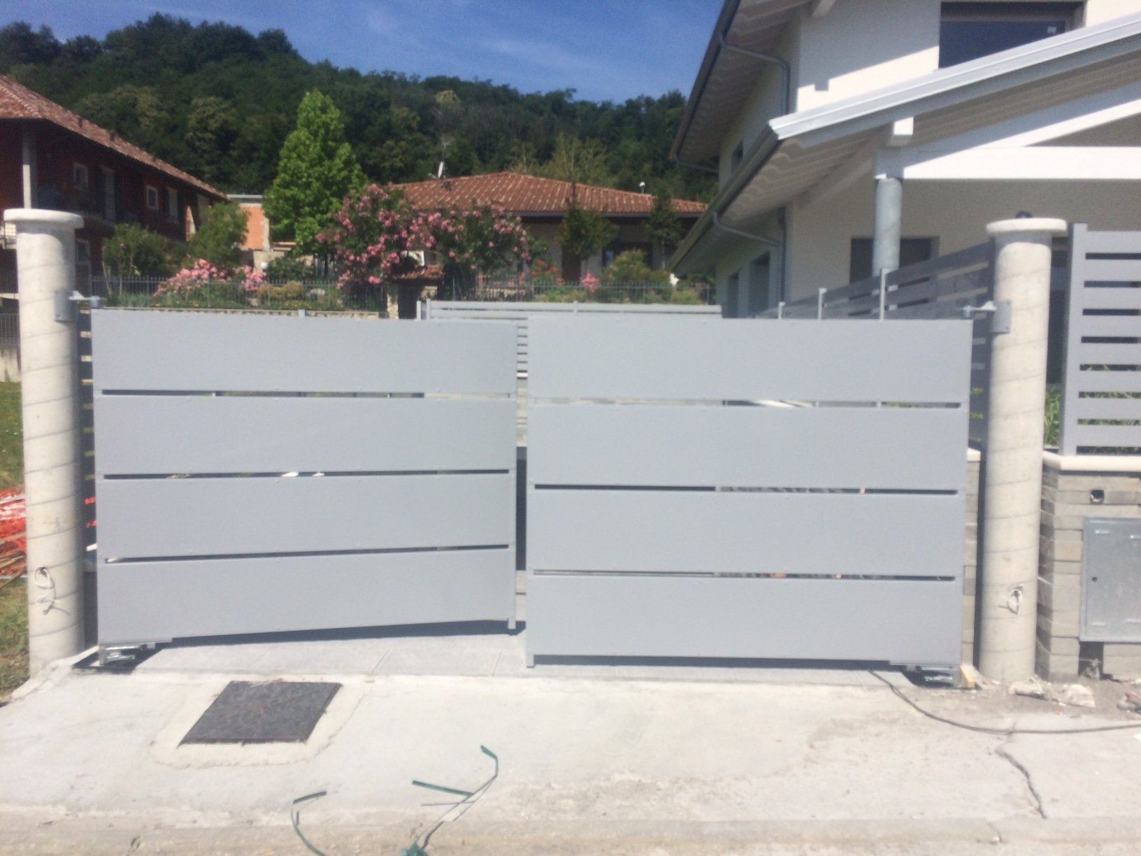 Cancello carrabile in metallo grigio con lamelle orizzontali davanti a una casa e al giardino, all'aperto in una giornata di sole.
