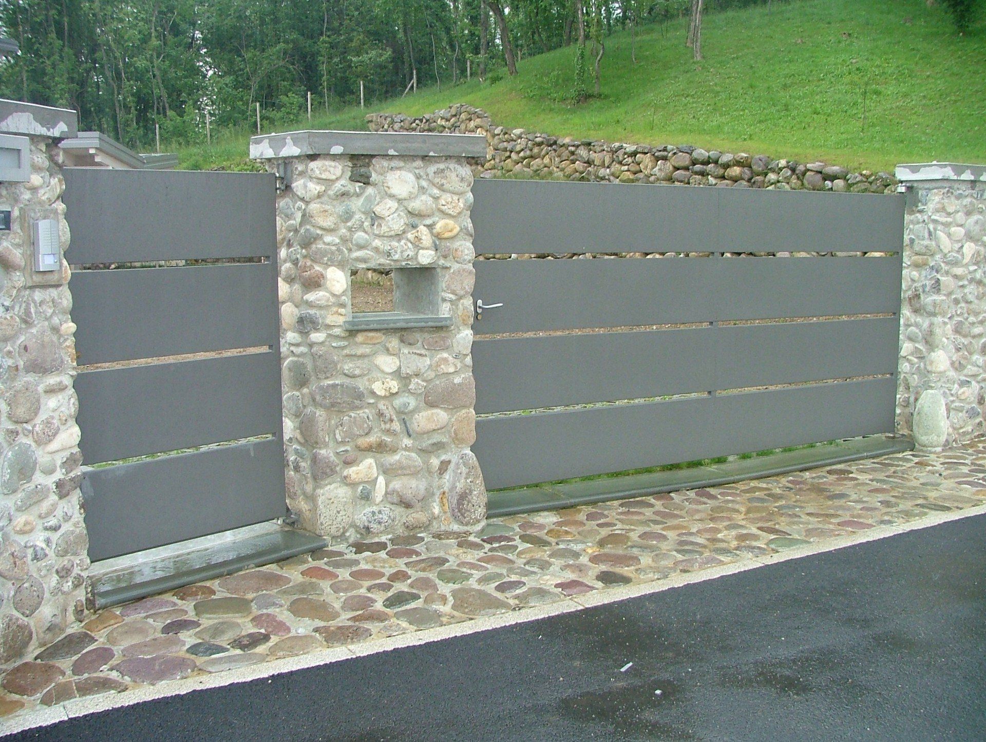 Cancello a doghe grigie con pilastri in pietra accanto a un vialetto pavimentato e a una collina erbosa.