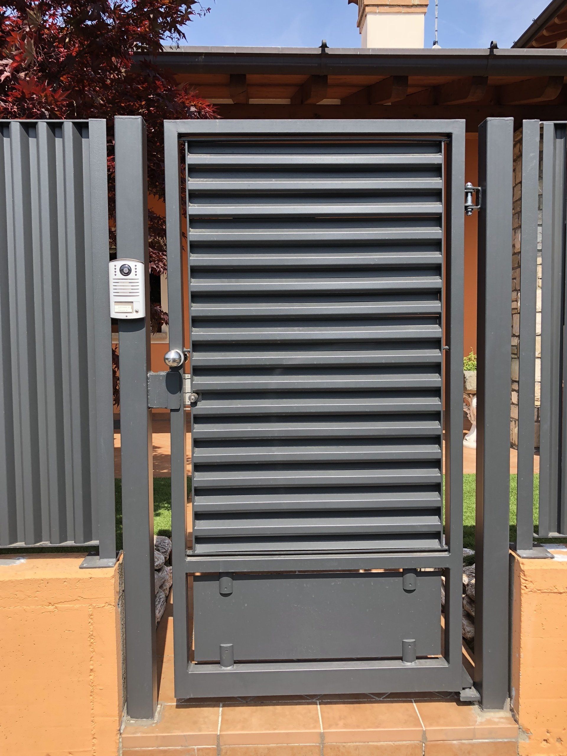 Cancello pedonale in metallo grigio con doghe orizzontali, integrato in una recinzione, accanto a un pannello per campanello a muro.