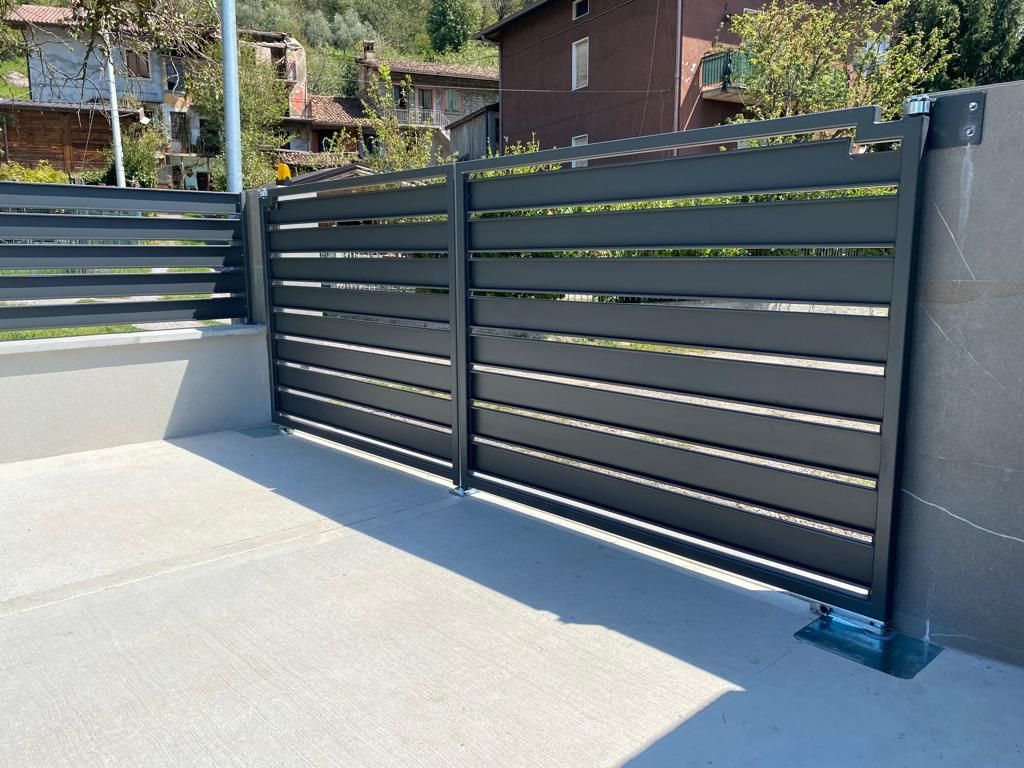 Recinzione e cancello in metallo nero a listelli orizzontali lungo un vialetto grigio chiaro in un soleggiato giardino residenziale.