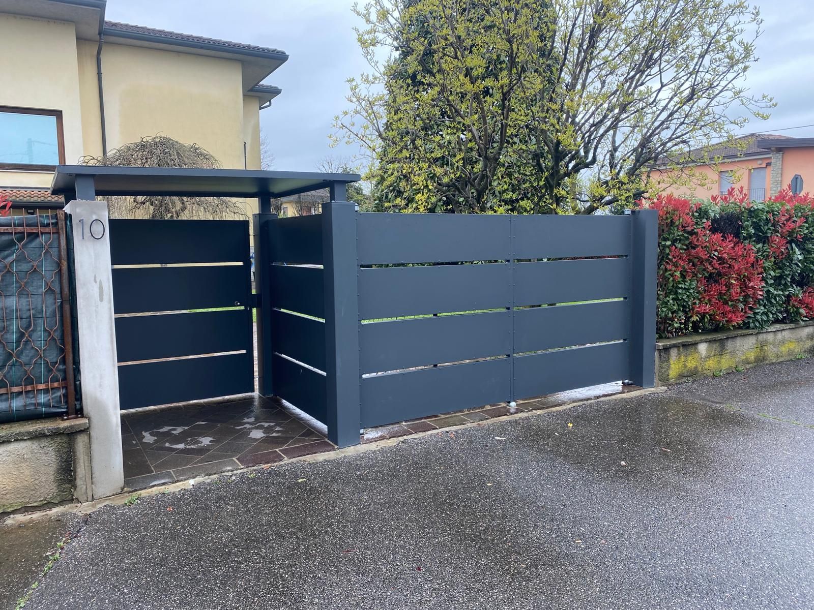 Cancello scorrevole moderno di colore grigio e cancello pedonale in un vialetto di accesso residenziale adiacente a una casa e al giardino.