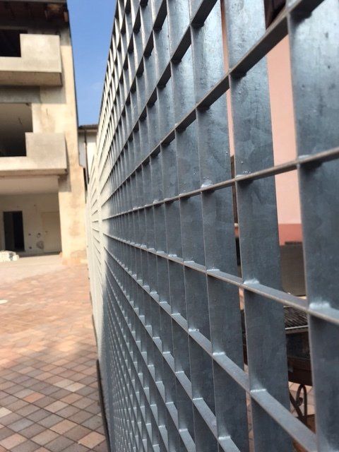 Recinzione a griglia metallica accanto a un cortile in mattoni e a una parete di un edificio beige.
