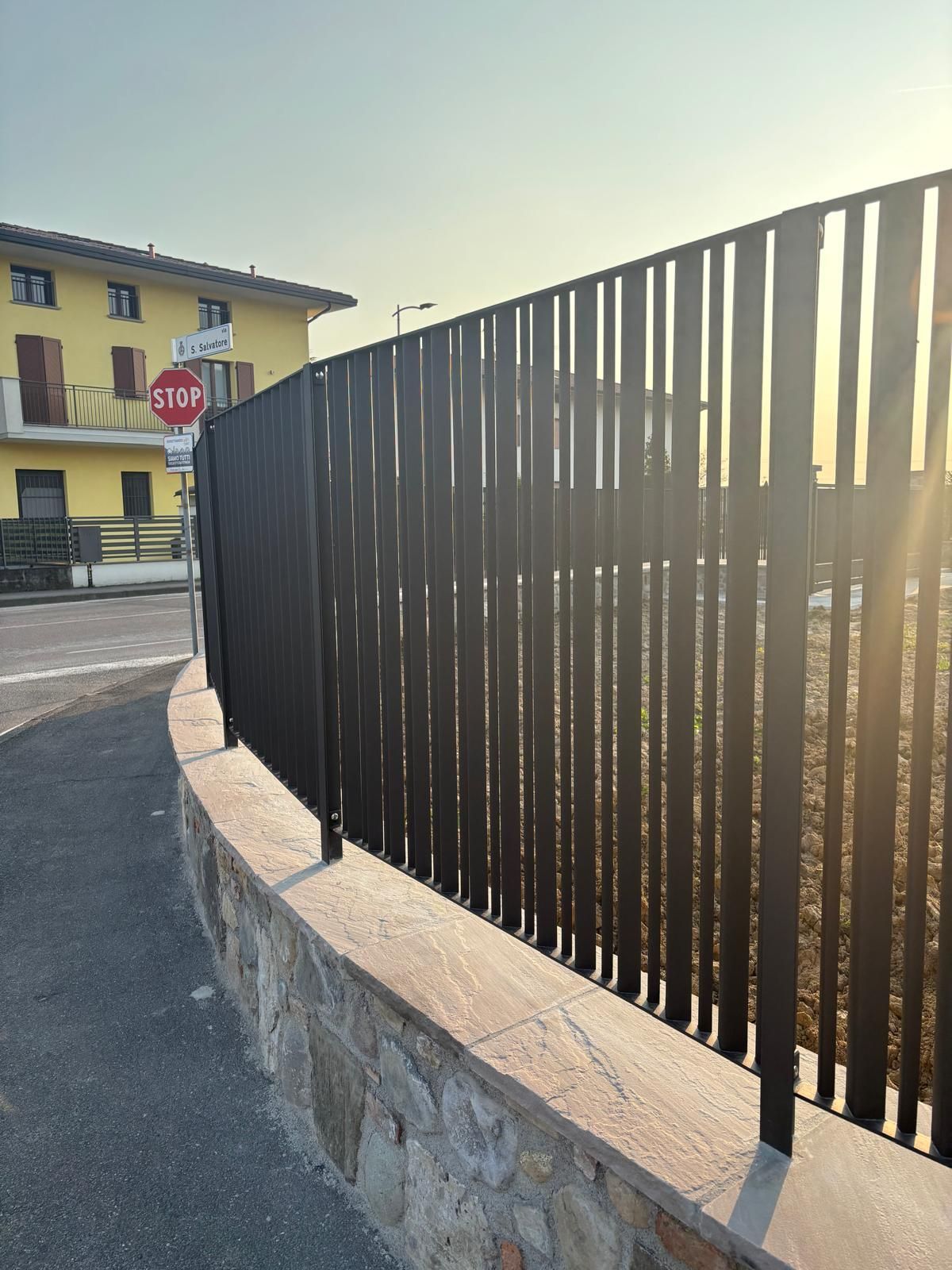 Muro di pietra curvo con un'alta recinzione metallica nera accanto a un edificio giallo, illuminato dal sole splendente.