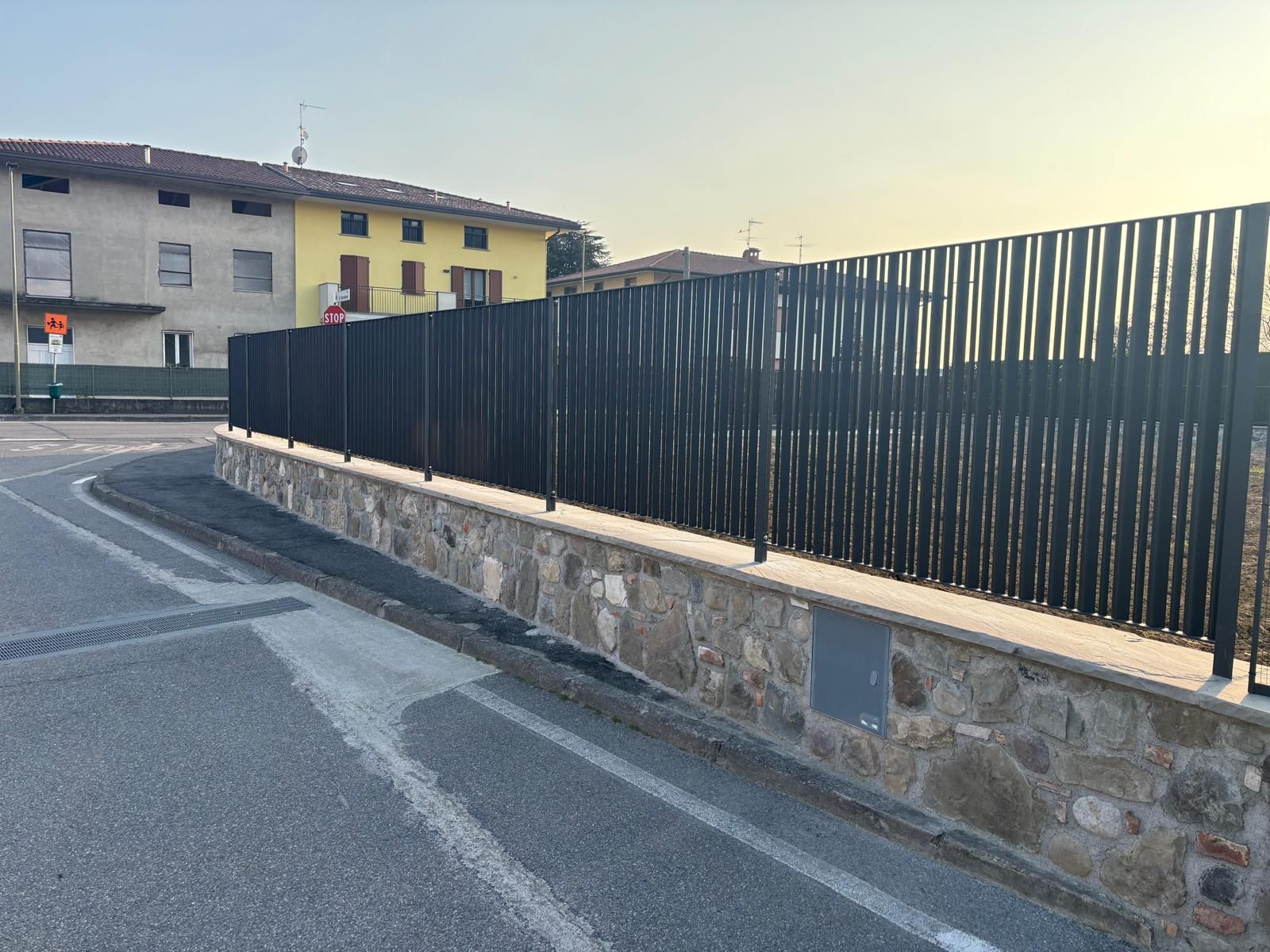 Una strada sinuosa costeggia un muro di pietra e una recinzione nera, con le case sullo sfondo al tramonto.