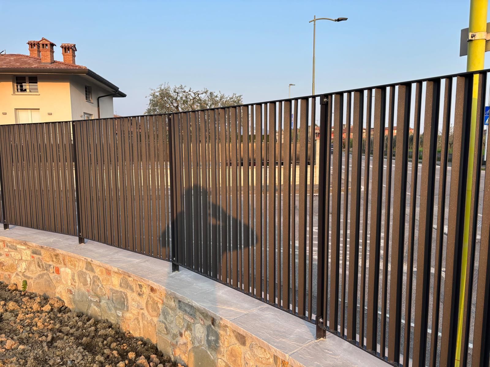 Recinzione metallica scura lungo un muro di contenimento in pietra a lato di una strada residenziale.