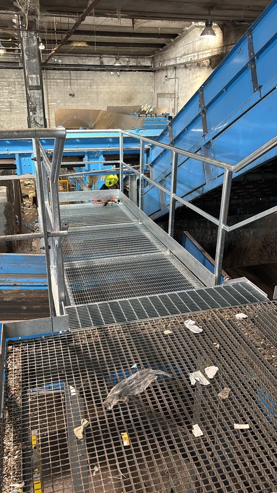 Passerella industriale con grigliato metallico, scale blu e ringhiere all'interno di una struttura simile a un magazzino.