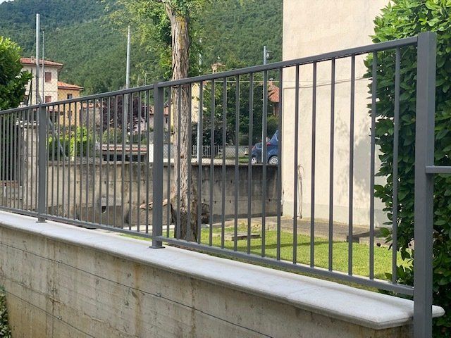 Recinzione metallica nera su un muro di cemento accanto a un edificio, con alberi e colline sullo sfondo.