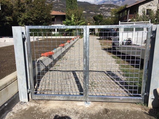Un cancello metallico chiuso blocca un vialetto di ghiaia, con case e montagne sullo sfondo.