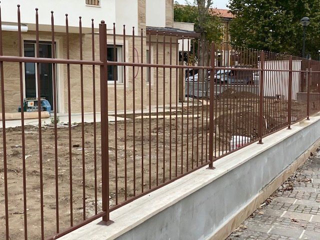 Recinzione metallica marrone lungo un basso muro di cemento accanto a un marciapiede e al cortile di un edificio.