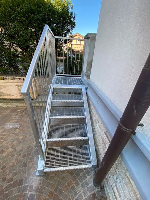 Scala esterna in metallo con corrimano accanto a un edificio bianco e a un vialetto in mattoni.