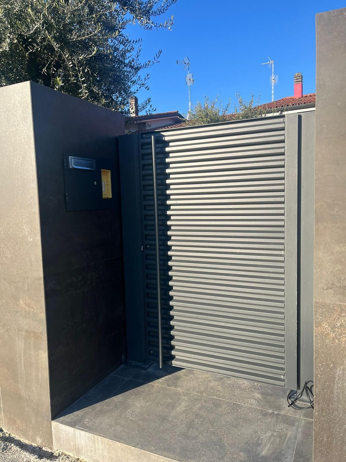 Cancello metallico grigio accanto a un muro d'ingresso scuro sotto un cielo azzurro