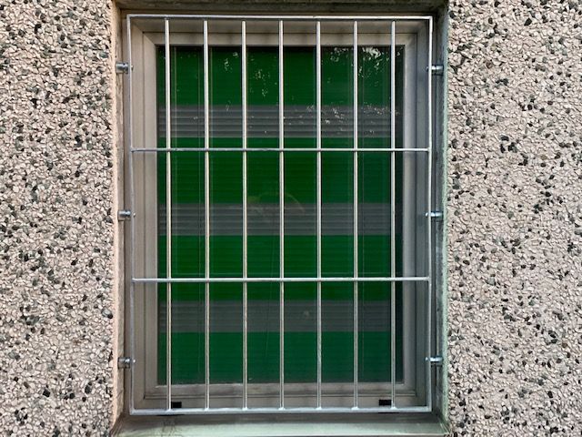Piccola finestra con sbarre e persiane verdi incastonata in un muro di cemento screziato.
