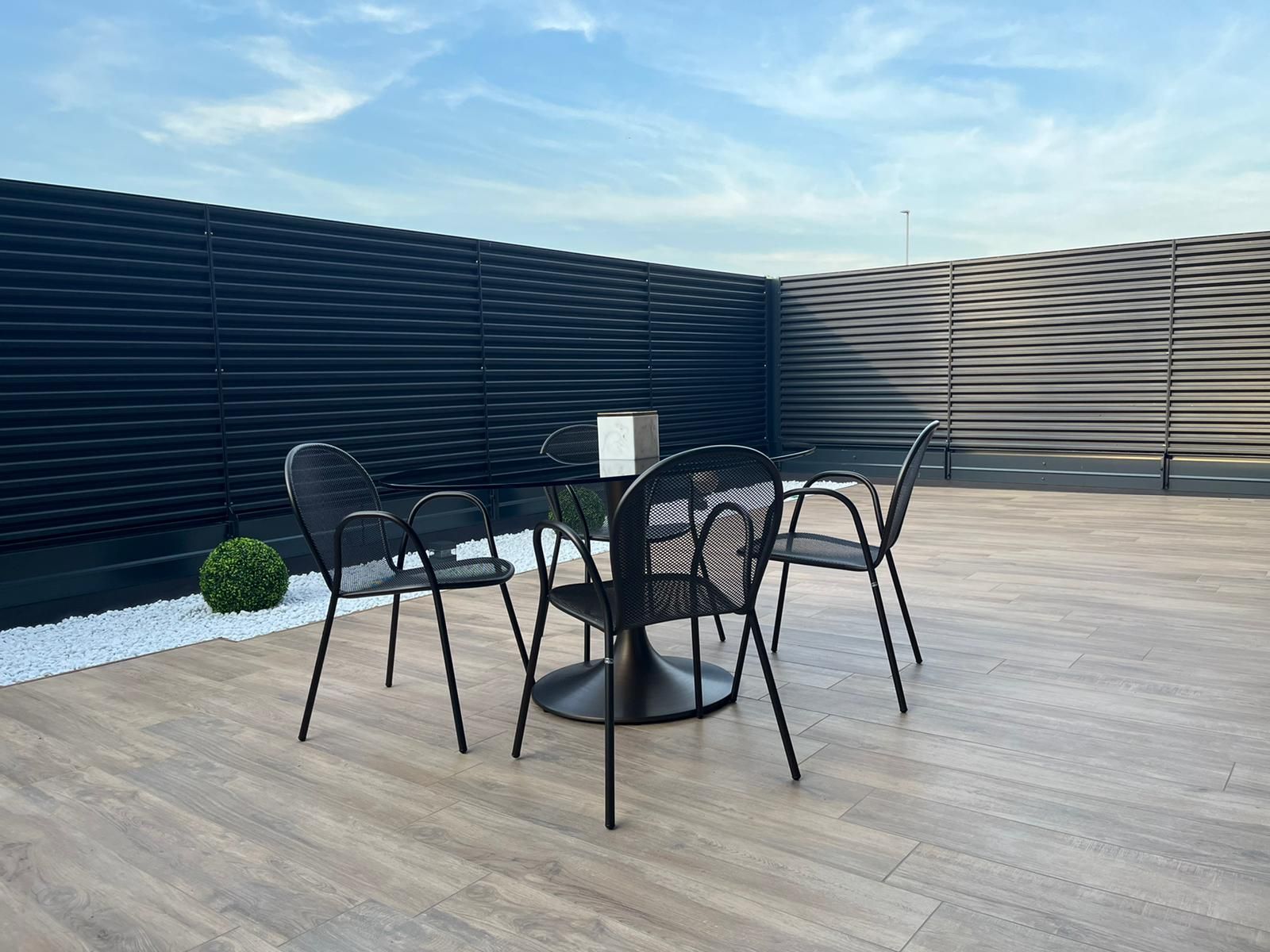 Patio con tavolo e sedie in metallo nero su una terrazza in legno chiaro, circondato da una recinzione scura che garantisce la privacy.