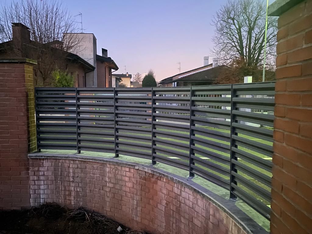 Recinzione curva a listelli grigio scuro lungo un muro di mattoni nel cortile di un'abitazione al crepuscolo.