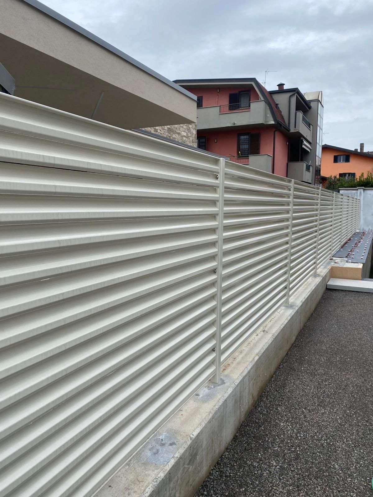 Recinzione ondulata bianca lungo un muro di cemento accanto a una strada residenziale con case sullo sfondo.