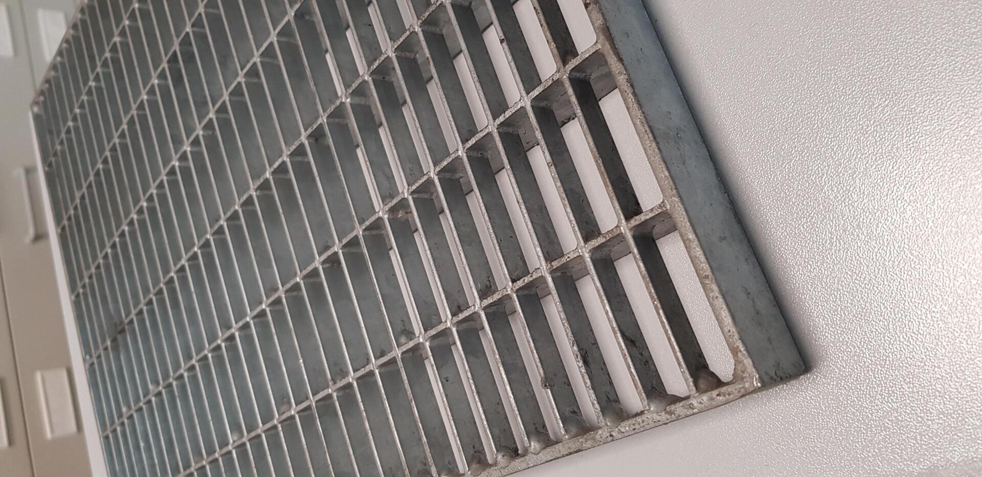Primo piano di una griglia di ventilazione a muro con griglie metalliche grigie su una parete bianca punteggiata.