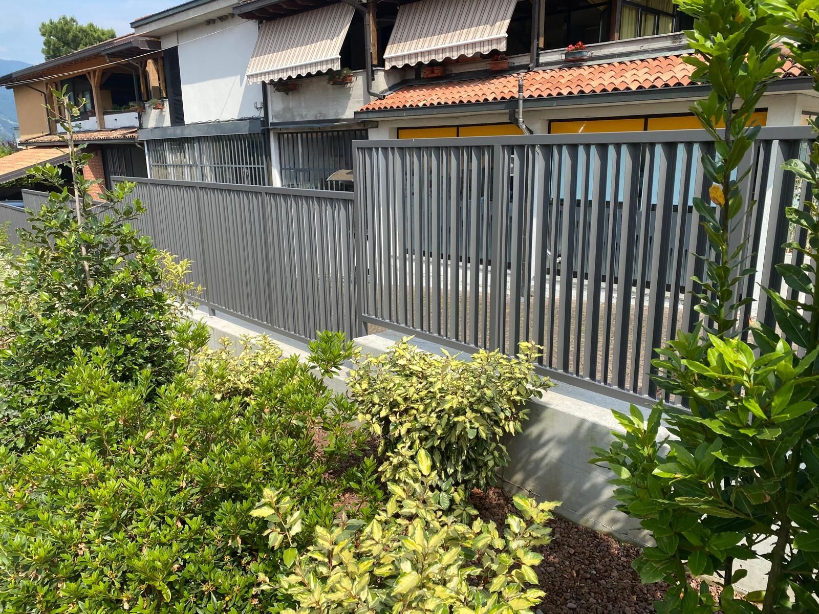 Casa residenziale con recinzione metallica grigia e cespugli verdi in primo piano.