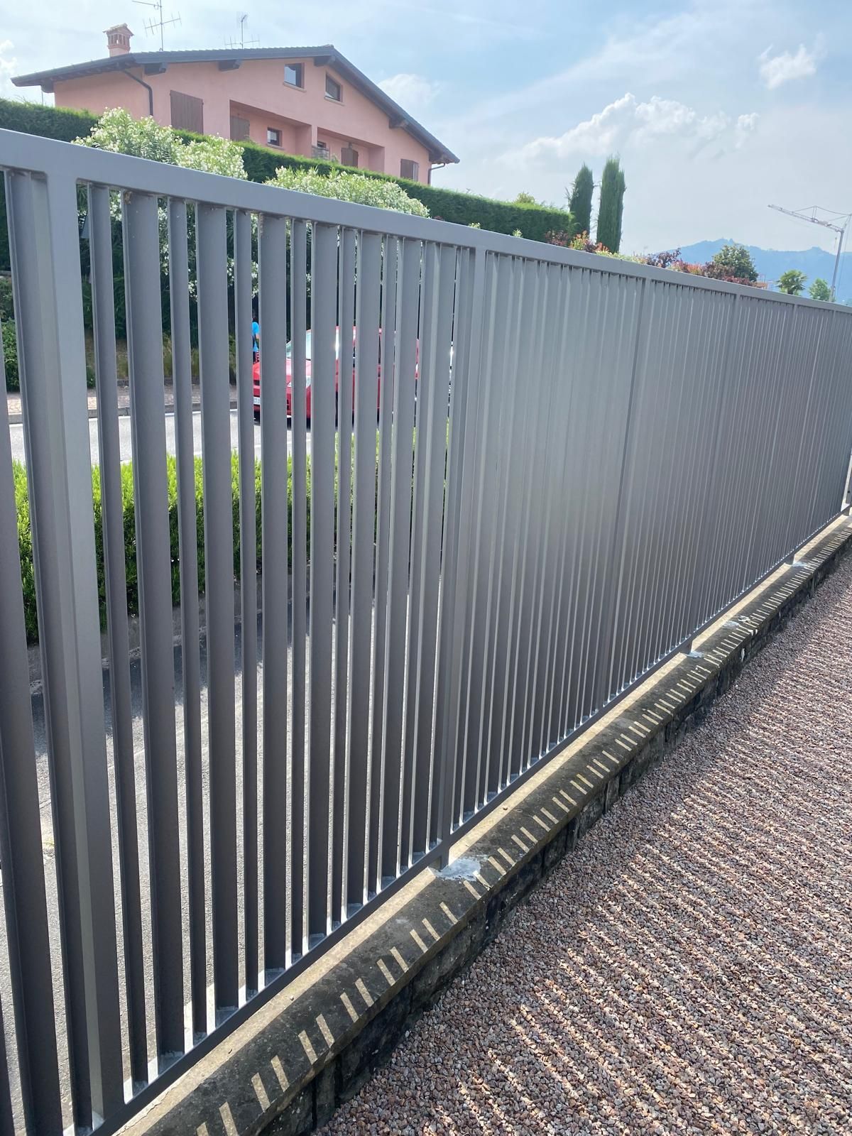Una lunga recinzione metallica grigia costeggia un marciapiede a lato di una strada, con case e alberi sullo sfondo.