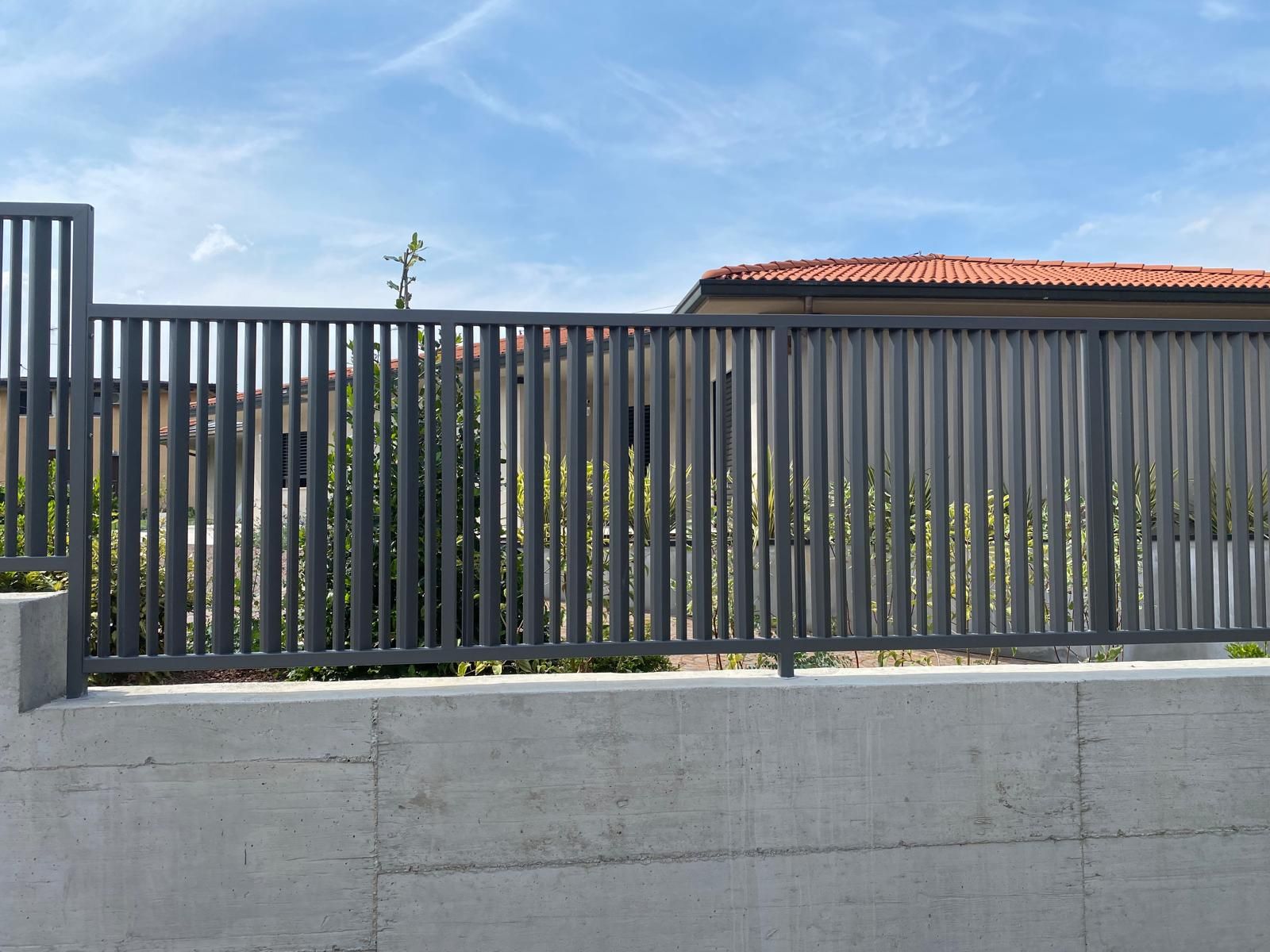 Una moderna recinzione metallica davanti a un muro di cemento e a una casa sotto un cielo azzurro.
