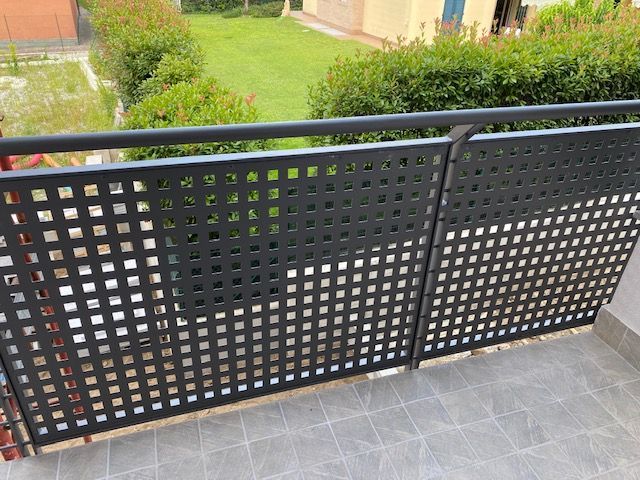 Ringhiera del balcone a traliccio nera accanto a un pavimento piastrellato grigio, con siepi verdi sullo sfondo.