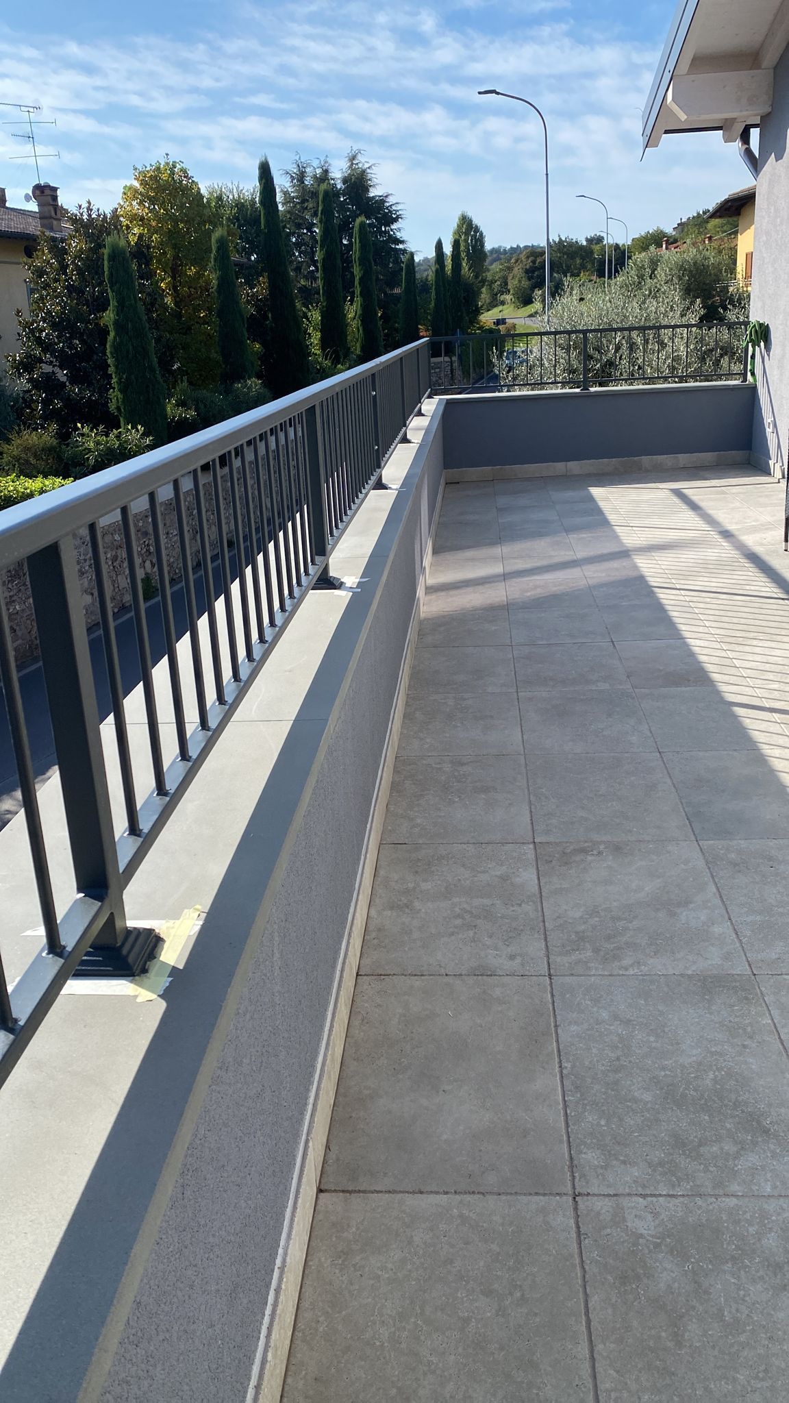 Balcone soleggiato con pavimento piastrellato in grigio, ringhiera scura e alberi sullo sfondo.