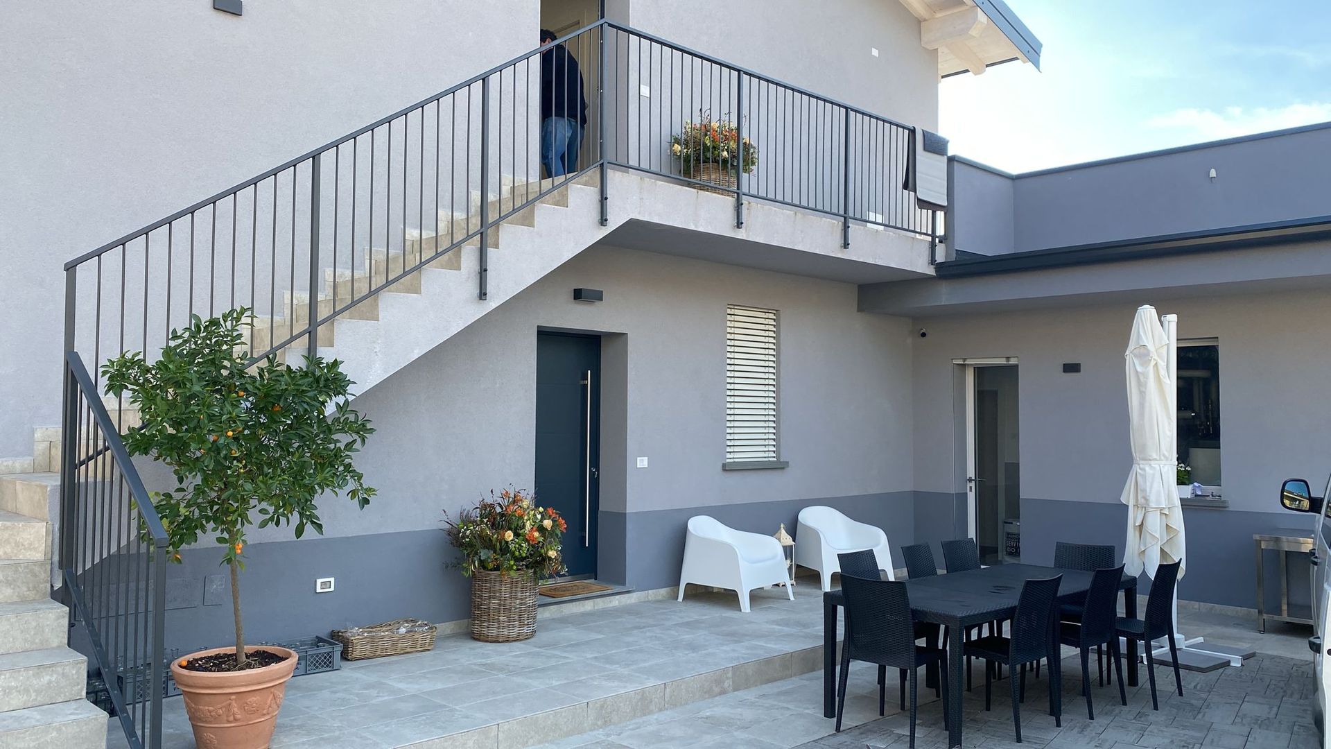 Cortile con patio, tavolo da pranzo all'aperto, sedie a sdraio, piante in vaso e scala esterna che conduce a un balcone superiore.