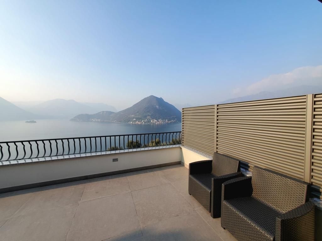 Terrazza con vista lago, due sedie nere, un paravento beige e montagne avvolte nella foschia in lontananza.