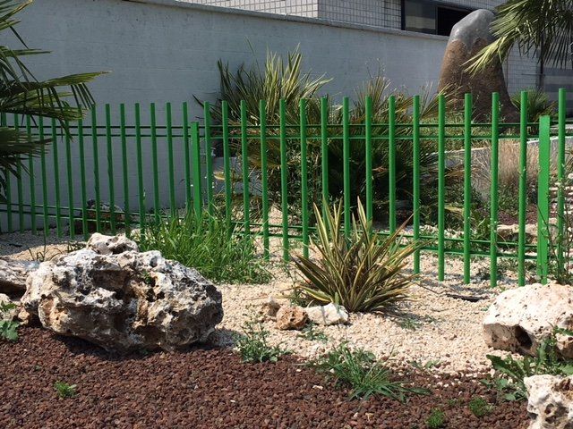 Recinzione metallica verde accanto a un paesaggio desertico con rocce, ghiaia e piante spinose vicino a un edificio