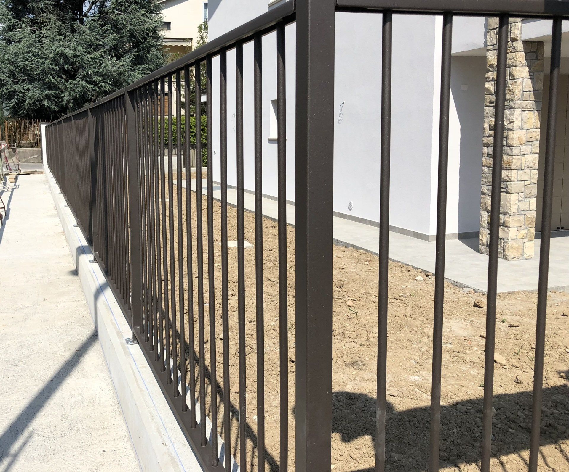 Una recinzione metallica nera lungo un marciapiede accanto a un muro bianco e a un cortile di terra in una giornata di sole.