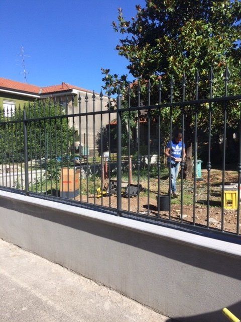Una persona con una camicia blu lavora in un giardino recintato accanto a un edificio in una giornata di sole.