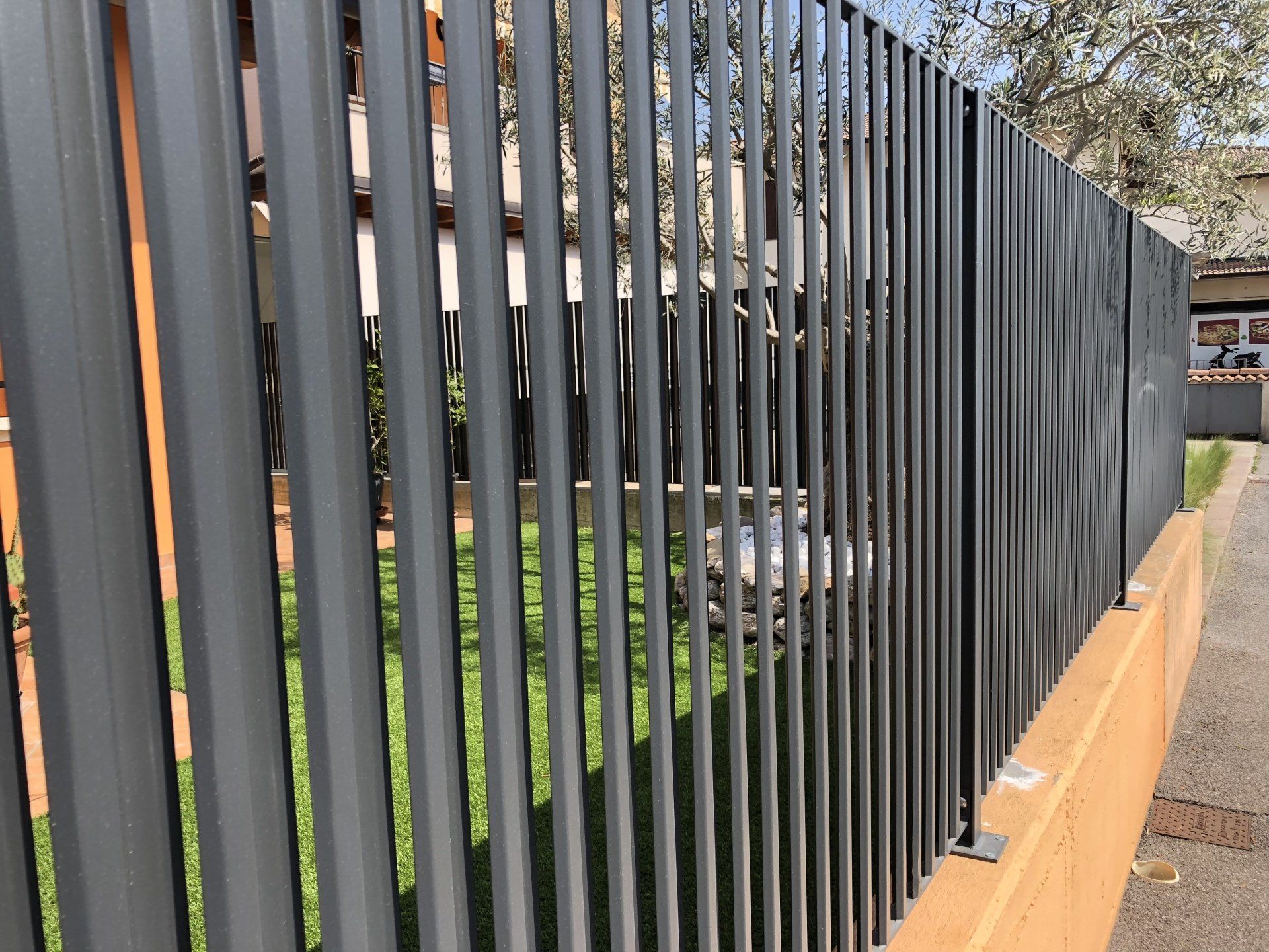 Una recinzione metallica grigio scuro costeggia un marciapiede, con un prato e un edificio alle sue spalle.