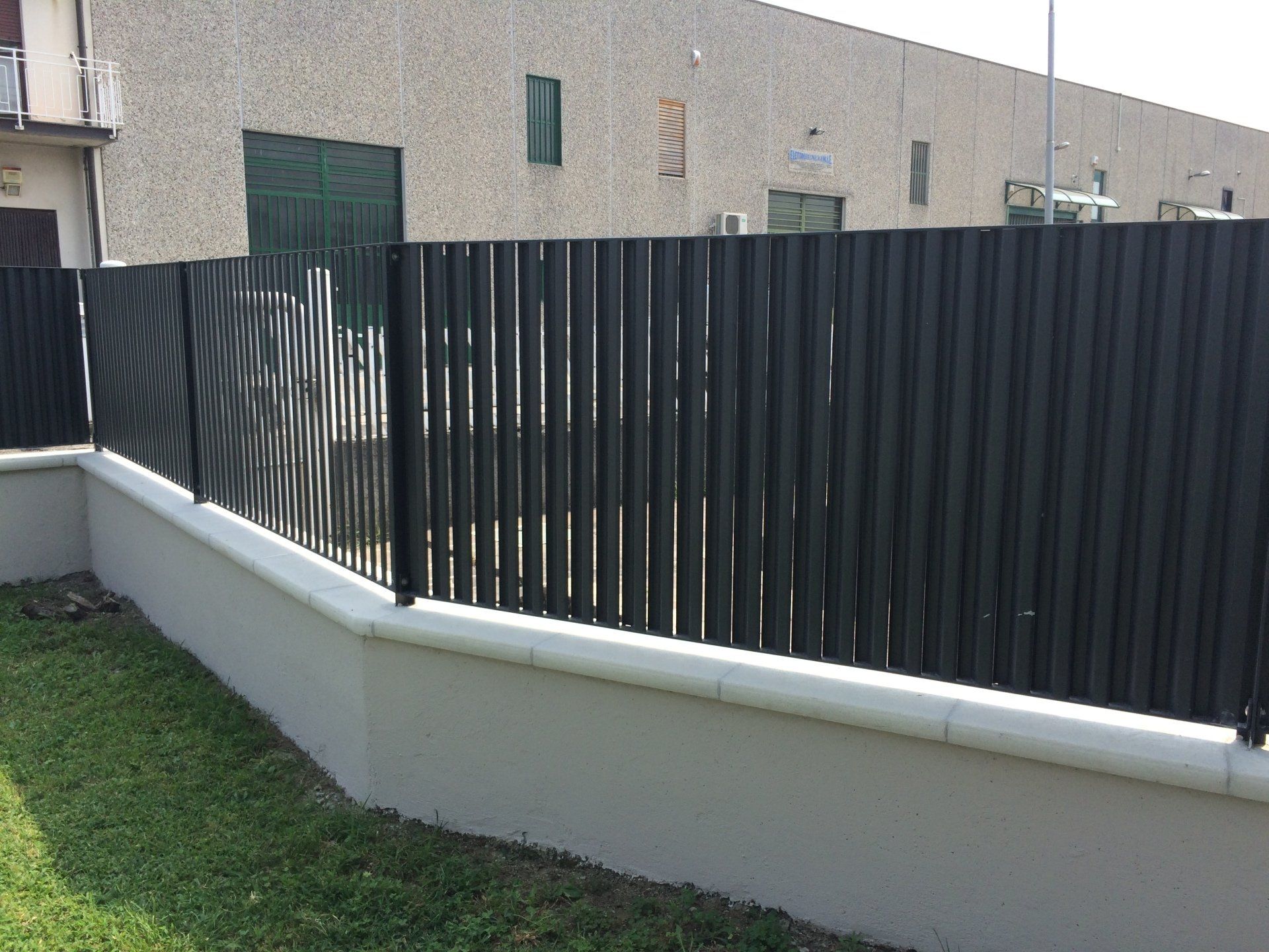 Muro di contenimento bianco con recinzione verticale scura lungo un prato accanto a un edificio beige.