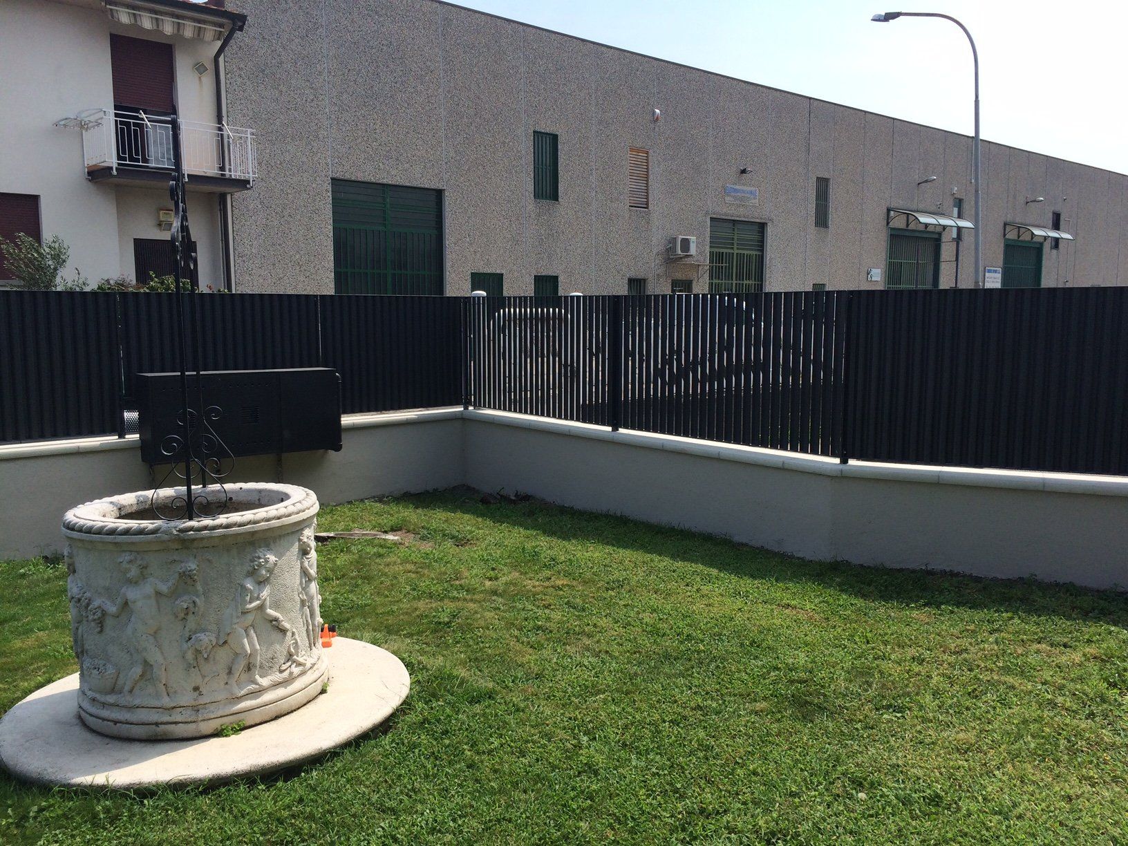 Piccolo cortile erboso con una fioriera cilindrica bianca e una recinzione nera accanto a edifici beige.