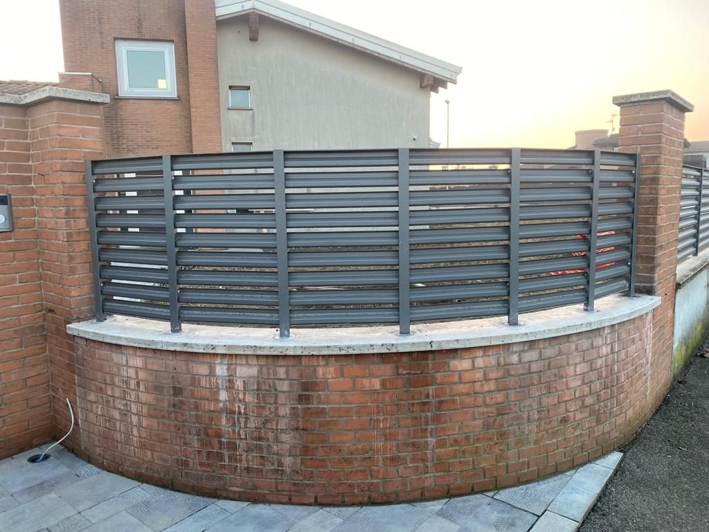 Muro di mattoni curvo con recinzione metallica a doghe grigie davanti a una casa al tramonto