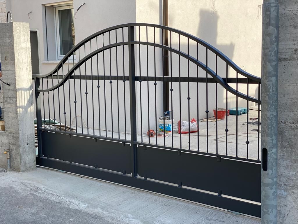 Cancello carrabile in metallo nero con parte superiore ad arco, posto di fronte a un muro e un marciapiede beige.