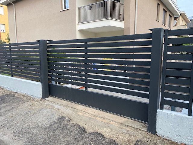 Moderna recinzione a doghe grigio scuro con pali in cemento di fronte a un condominio beige.