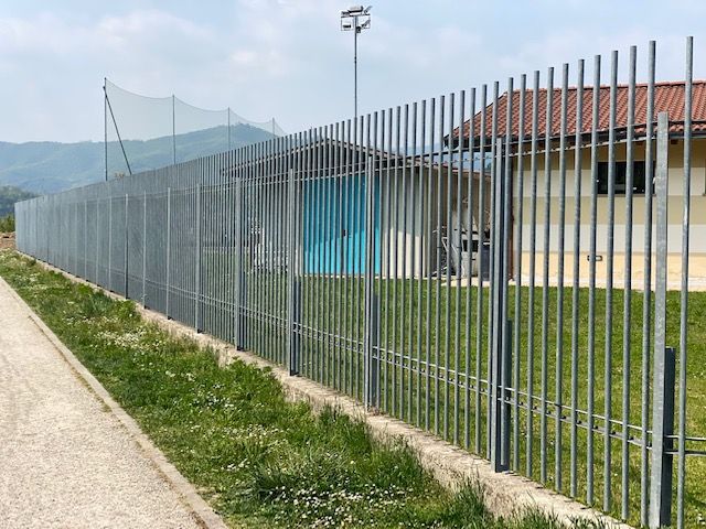 Una lunga recinzione metallica costeggia un sentiero, con un edificio e le montagne sullo sfondo.