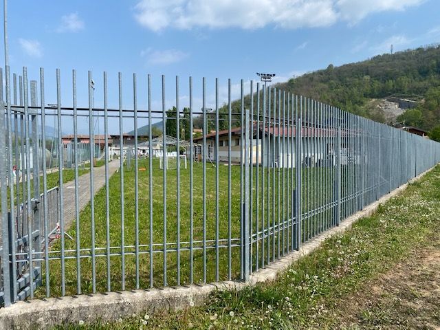Recinzione metallica attorno a un'area erbosa a lato di una strada, con un ponte e una collina sullo sfondo.