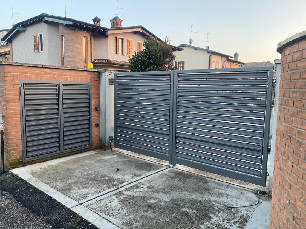 Cancello carrabile e cancello laterale in metallo grigio accanto a un muro di mattoni, con case visibili sullo sfondo.