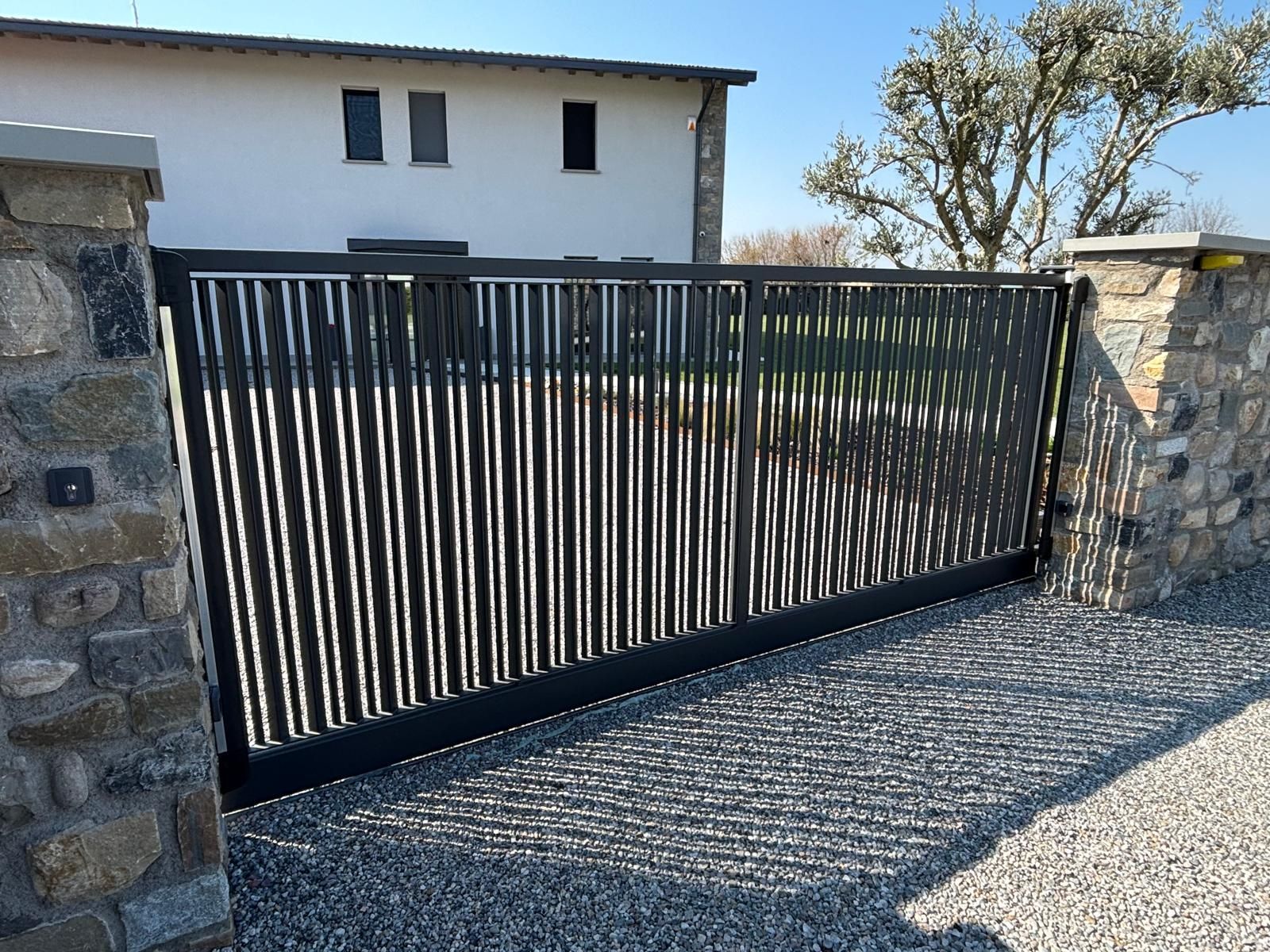 Cancello d'ingresso in metallo nero tra pilastri di pietra di fronte a una casa bianca.