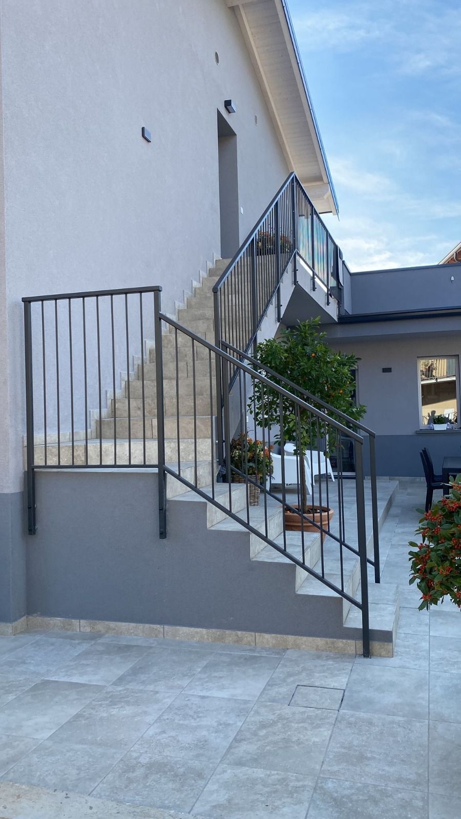 Scala esterna con ringhiera grigia accanto a una casa bianca e patio piastrellato.