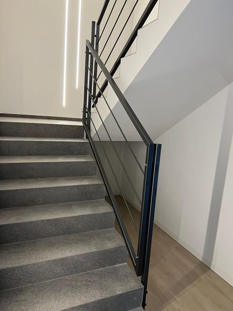 Vano scale interno con ringhiera in metallo nero e gradini rivestiti in moquette grigia, accanto a un pavimento in legno chiaro.