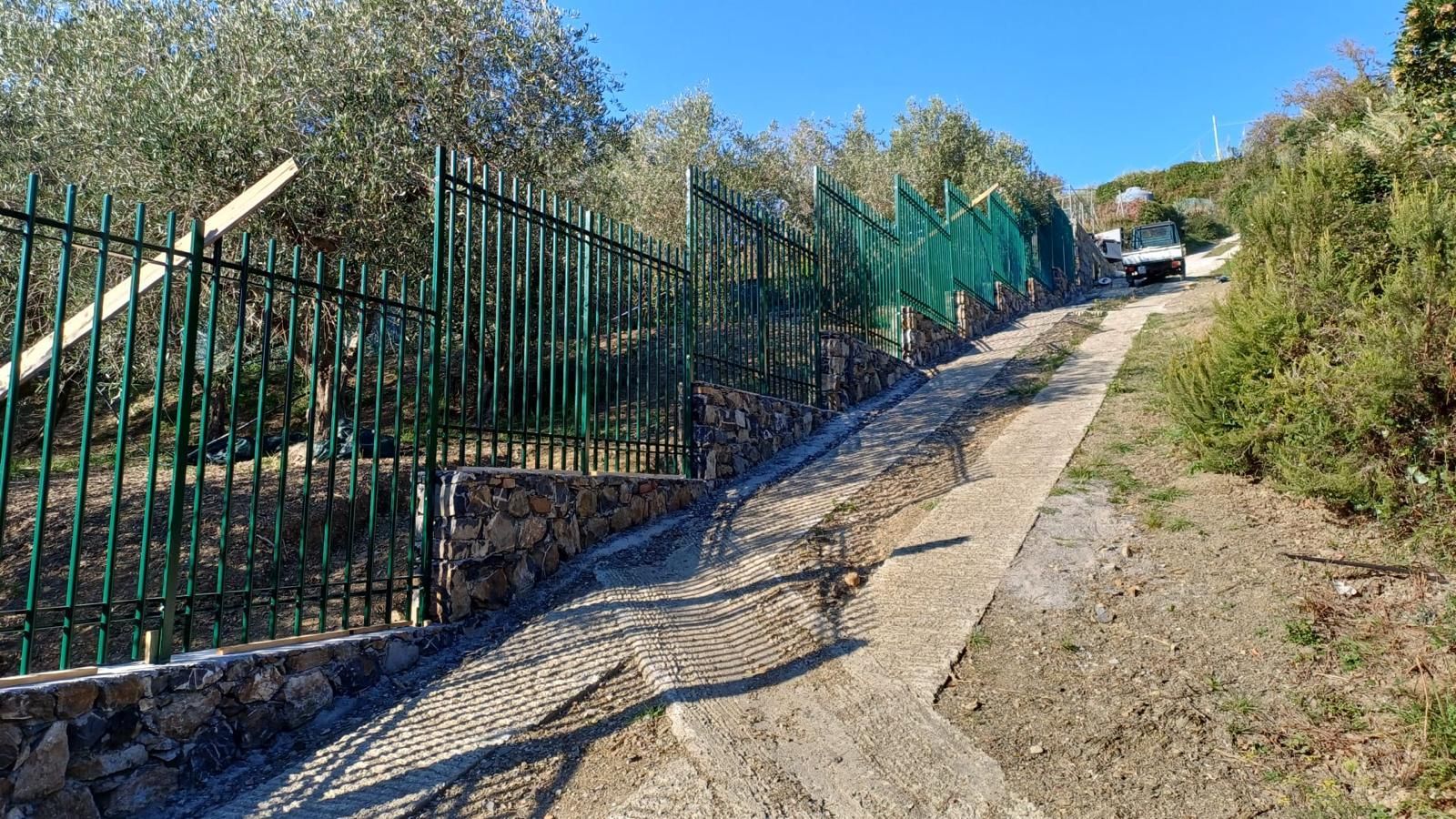 Un ripido sentiero sterrato costeggia una recinzione verde, con cespugli e un'auto parcheggiata in cima, sotto un cielo azzurro.