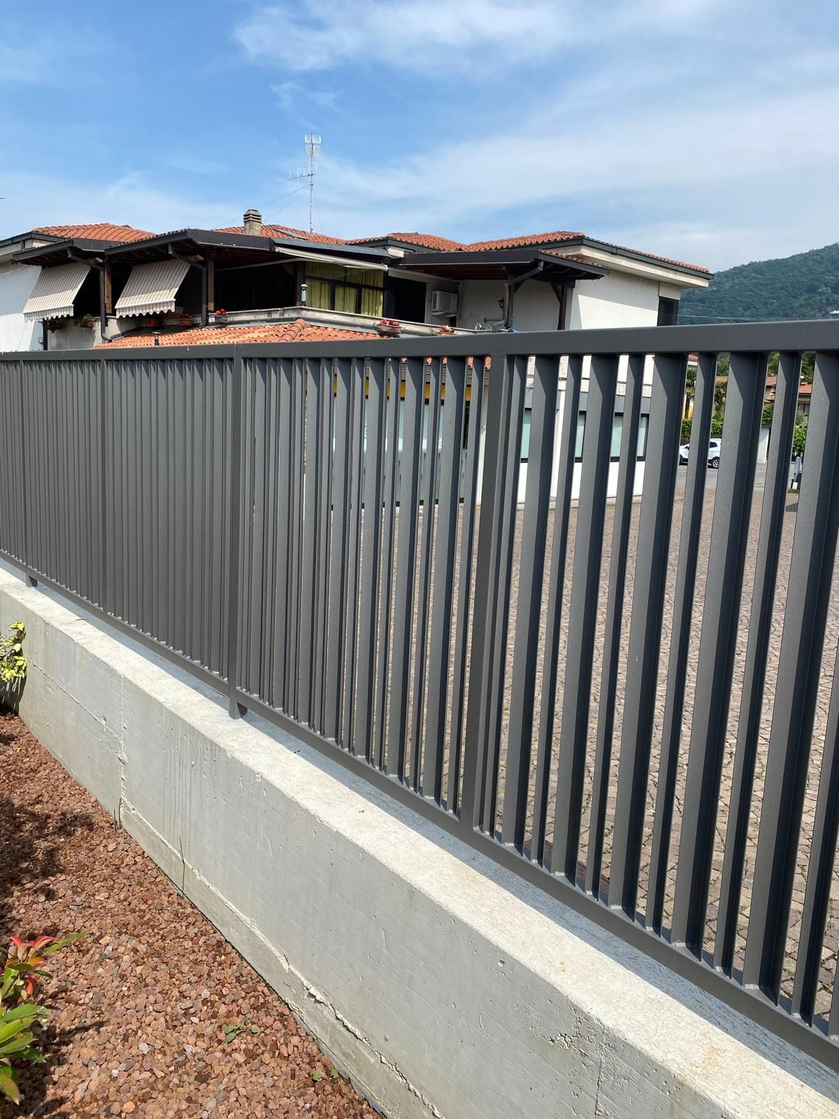 Recinzione metallica accanto a un edificio con il tetto danneggiato, sotto un cielo sereno, con un muro di cemento e la strada in primo piano.